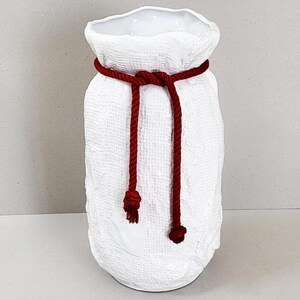 Può includere: Un vaso in ceramica bianca con una superficie testurizzata che ricorda un sacco di tessuto. Il vaso presenta una corda rosso scuro annodata attorno al collo, con due estremità che pendono sul davanti, ciascuna terminante con una piccola nappa.