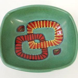 May include: A green ceramic dish with a red and yellow geometric design. The design is a stylized shape resembling a figure eight.