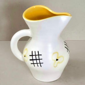 Puede incluir: Una jarra de cerámica blanca con acabado brillante, que presenta un borde interior y un asa de color amarillo intenso. El exterior está decorado con patrones de cuadrícula negros pintados a mano y formas florales abstractas amarillo pálido.