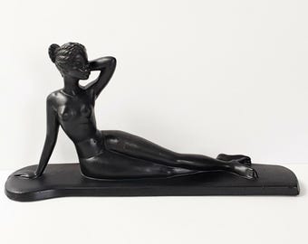 Può includere: Una statuetta nero opaco raffigurante una persona sdraiata che poggia su un gomito, posizionata su una base allungata abbinata. La figura ha una finitura liscia, con un braccio sollevato dietro la testa e le gambe distese orizzontalmente.