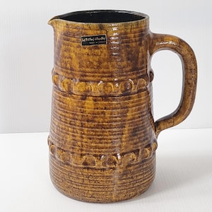 以下が含まれることがあります： テクスチャ加工された表面と大きなハンドルが付いた茶色のセラミックピッチャー。ピッチャーは内側が暗く、円形のアクセントが付いた装飾的な水平バンドがあります。「Les Potiers d'Aiguiry Made in France」と書かれたラベルが付いています。