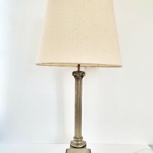 Op de afbeelding: Een tafellamp met een hoge, zilverkleurige metalen voet in de vorm van een gecanneleerde zuil met een vierkante sokkel. De lamp is voorzien van een grote, taps toelopende, gebroken witte stoffen lampenkap met een subtiele textuur en rand.