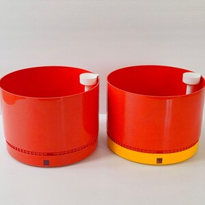 May include: Two cylindrical planters with a bright red exterior and a white interior. Each planter features a small white spout on the inner rim and a perforated base section. One planter has a solid red base, while the other has a yellow base.