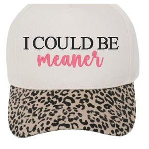 May include: A white baseball cap with the text "I COULD BE meaner" in black and pink. The brim features a tan and black leopard print pattern. This cap is a stylish accessory.