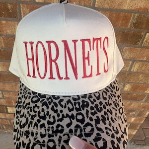 May include: A white baseball cap with the word "HORNETS" embroidered in red. The brim features a black and tan leopard print pattern. The cap is a classic, structured style.