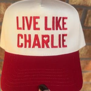 May include: A red and white baseball cap with the text "LIVE LIKE CHARLIE" embroidered in red. The cap has a red brim and a white crown, with a small red heart symbol in the text.