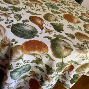 May include: A white tablecloth featuring a repeating pattern of pumpkins and gourds in shades of orange, dark green, and muted yellow, interspersed with green vines, leaves, and small flowers printed across the entire fabric surface.