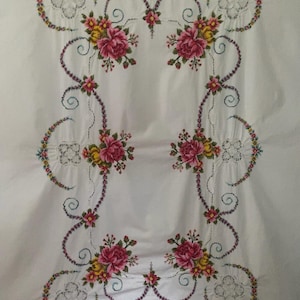 May include: A white rectangular textile featuring intricate cross-stitch embroidery of pink and yellow roses. The floral pattern is arranged in a symmetrical border with decorative scrollwork and openwork lace details along the edges.