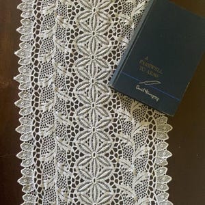 May include: A cream-colored lace table runner featuring an intricate floral and leaf pattern with scalloped edges, positioned next to a dark blue hardcover book titled A Farewell to Arms by Ernest Hemingway with a small feather graphic.
