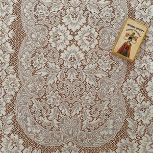 May include: A white lace fabric featuring an intricate floral and scrollwork pattern with a mesh background. A small rectangular book titled Madame Bovary by Gustave Flaubert with a yellow border and a graphic illustration is placed on the lace.