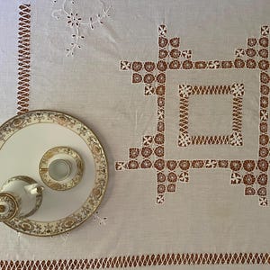 May include: A light beige linen tablecloth featuring intricate brown cutwork embroidery in a geometric square pattern, accompanied by a white porcelain tea set with gold filigree trim, including a small teapot, a saucer, and a teacup.