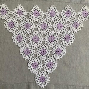 May include: A triangular crochet shawl featuring a repeating pattern of small, light purple square motifs connected by an intricate white lace border. The design consists of multiple lavender-colored centers surrounded by white yarn loops.
