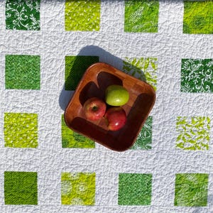 May include: A square wooden bowl holds three apples, two red and one green, on a white quilt with green square patterns. The quilt has various green fabric patterns, including floral and fern designs.