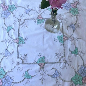 May include: A white tablecloth featuring intricate embroidery in shades of light blue, mint green, and soft pink, arranged in a square pattern. A clear glass vase containing a cluster of vibrant purple flowers sits atop the embroidered fabric.