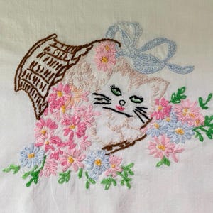 May include: A white fabric panel featuring an embroidered design of a cat peeking out from a brown woven basket. The cat is surrounded by pink and light blue flowers with green stems and leaves. A light blue ribbon bow is tied above the cat.