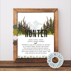 May include: A framed print with a rustic wooden frame. The artwork features the word "HUNTER" in bold letters, with a watercolor image of antlers, trees, and mountains. Text below describes the meaning of the name. A circular sticker with the text "MAKES MY DAY SHOP HAPPY" is in the bottom right corner.