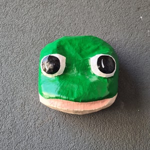 Op de afbeelding: Een handgemaakt, groen kikkergezicht met grote zwarte ogen en een lichtroze mond. Het gezicht van de kikker is geschilderd op een houten blok, met een grillig en speels ontwerp. Het kikkergezicht is een decoratief item.