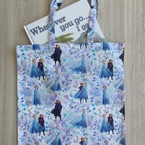 Op de afbeelding: Een witte canvas tote bag met een herhalend patroon van Frozen-karakters, waaronder Elsa en Anna, op een lichtblauwe achtergrond met paarse bloemen. De tas heeft een boek erin met de tekst "Wherever you go... I go".