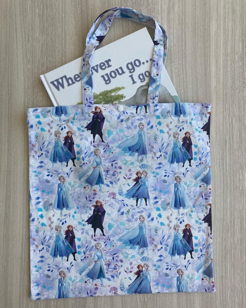Op de afbeelding: Een witte canvas tote bag met een herhalend patroon van Frozen-karakters, waaronder Elsa en Anna, op een lichtblauwe achtergrond met paarse bloemen. De tas heeft een boek erin met de tekst "Wherever you go... I go".