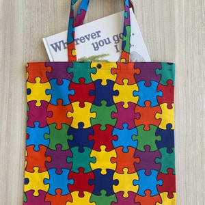 Op de afbeelding: Een kleurrijke tote bag met een puzzelstukjespatroon. De tas heeft een boek erin met de tekst "Wherever you go... I'll be there".