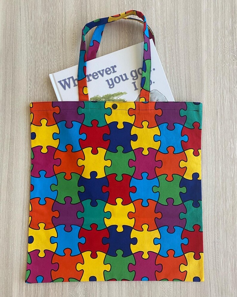 Op de afbeelding: Een kleurrijke tote bag met een puzzelstukjespatroon. De tas heeft een boek erin met de tekst "Wherever you go... I'll be there".