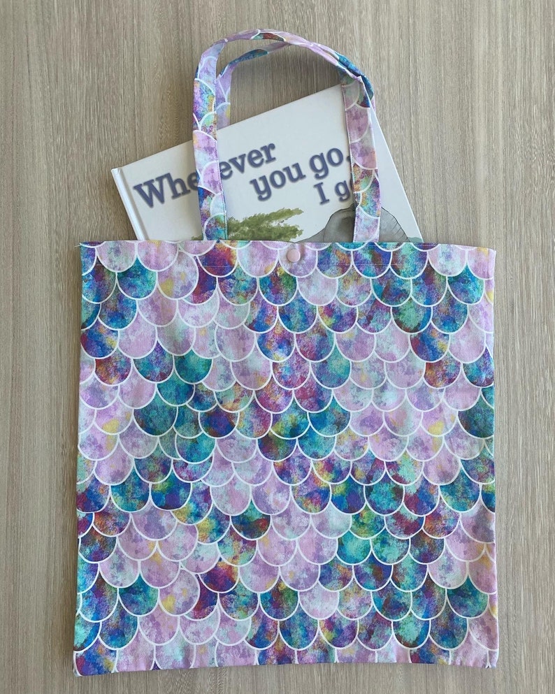 Op de afbeelding: Een herbruikbare draagtas met een zeemeermin-schubbenpatroon in tinten blauw, groen, paars en roze. De tas heeft een witte achtergrond en een enkele knoopsluiting. De tas wordt getoond met een boek erin dat "Wherever you go I go" zegt.