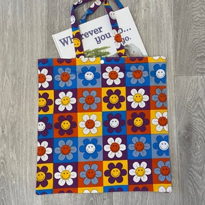 Op de afbeelding: Een paarse tote bag met een patroon van witte, gele, oranje en blauwe smiley-gezichten in bloemen. De tas heeft een boek erin dat zegt "Wherever you go..."