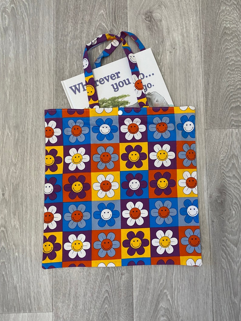 Op de afbeelding: Een paarse tote bag met een patroon van witte, gele, oranje en blauwe smiley-gezichten in bloemen. De tas heeft een boek erin dat zegt "Wherever you go..."
