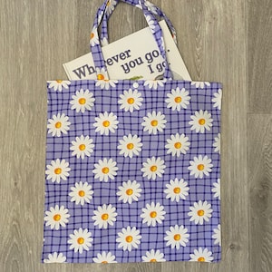 Op de afbeelding: Een paarse tote bag met een wit en geel madeliefjespatroon en een geruit ontwerp. De tas heeft een boek erin met de tekst "Wherever you go, I go."