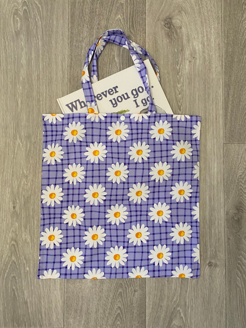 Op de afbeelding: Een paarse tote bag met een wit en geel madeliefjespatroon en een geruit ontwerp. De tas heeft een boek erin met de tekst "Wherever you go, I go."