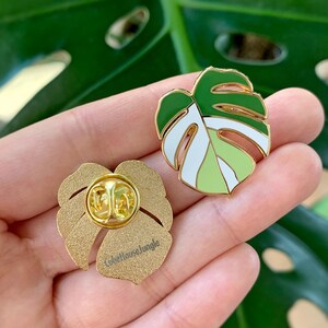 May include: Two gold-tone enamel pins featuring a green and white monstera leaf design. The back of one pin shows the text "Lovehousejungle".