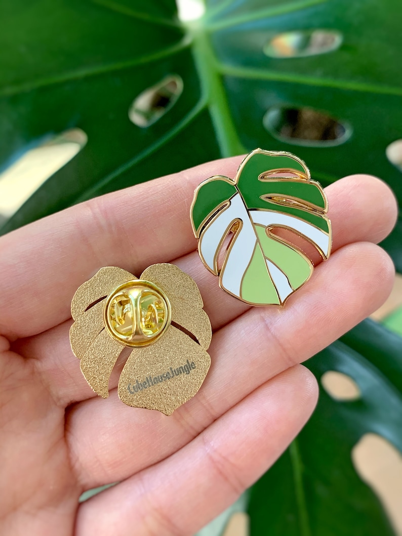 May include: Two gold-tone enamel pins featuring a green and white monstera leaf design. The back of one pin shows the text "Lovehousejungle".