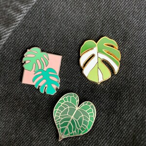 May include: Three enamel pins featuring different green leaf designs. The first pin is a pink square with two green monstera leaves. The second pin is a green and white monstera leaf. The third pin is a green heart-shaped leaf.