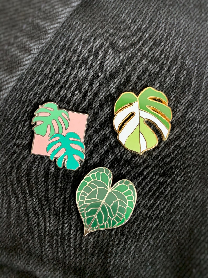May include: Three enamel pins featuring different green leaf designs. The first pin is a pink square with two green monstera leaves. The second pin is a green and white monstera leaf. The third pin is a green heart-shaped leaf.