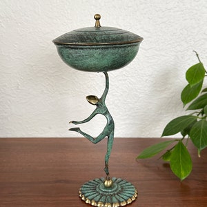 May include: A green bronze sculpture of a dancer holding a lidded dish. The dish has a green patina and a gold rim. The sculpture is on a round base with a gold rim.