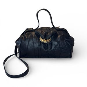 以下が含まれることがあります： ユニークなデザインの黒い革のハンドバッグ。バッグは、大きな金色のリングクラスプとステッチされたパッチワークパターンが特徴です。上部にハンドルと、取り外し可能な長いショルダーストラップが付いています。バッグは白い背景にあります。