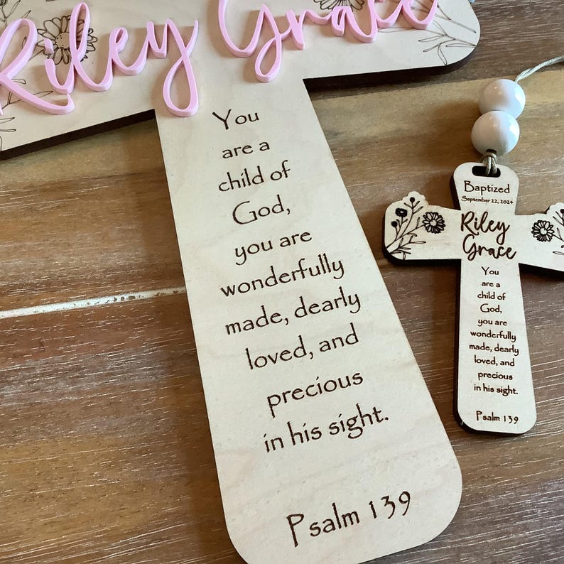 May include: A light-coloured wooden cross featuring the laser-engraved text "You are a child of God, you are wonderfully made, dearly loved, and precious in his sight. Psalm 139" and a smaller matching cross with white beads and the name "Riley Grace".