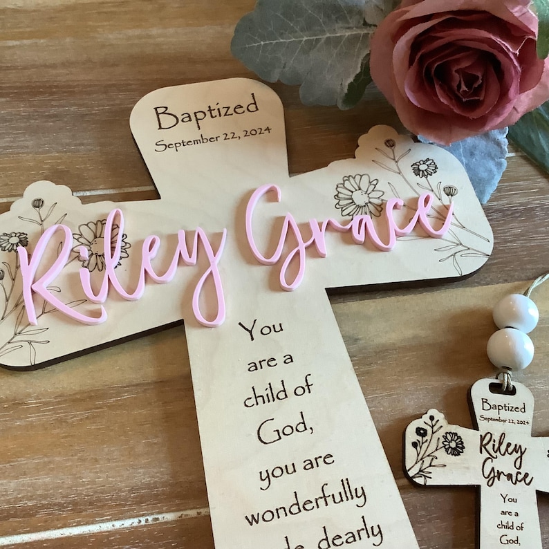 May include: A light-coloured wooden cross featuring the name Riley Grace in raised pink script lettering. The cross is engraved with the text Baptized September 22, 2024, floral line art, and the phrase You are a child of God, you are wonderfully made dearly.