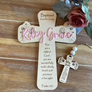 May include: A light-coloured wooden cross featuring the text "Baptized September 22, 2024", the name "Riley Grace" in raised pink script, and a verse from Psalm 139. A smaller matching wooden cross ornament with white beads is also included.