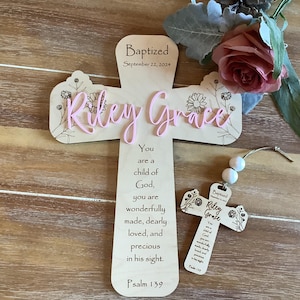May include: A light-coloured wooden cross featuring the text "Baptized September 22, 2024", "Riley Grace" in pink script, and a floral design. Below is the text "You are a child of God, you are wonderfully made, dearly loved, and precious in his sight. Psalm 139". A smaller matching cross ornament with two white beads is included.