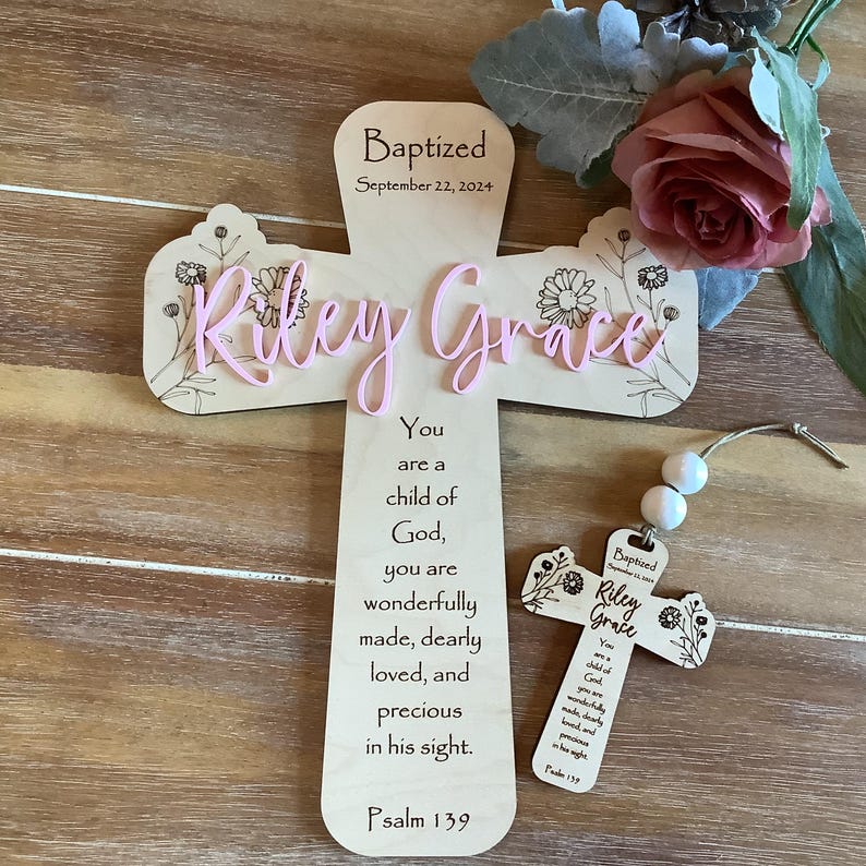 May include: A light-coloured wooden cross featuring the text "Baptized September 22, 2024", "Riley Grace" in pink script, and a floral design. Below is the text "You are a child of God, you are wonderfully made, dearly loved, and precious in his sight. Psalm 139". A smaller matching cross ornament with two white beads is included.