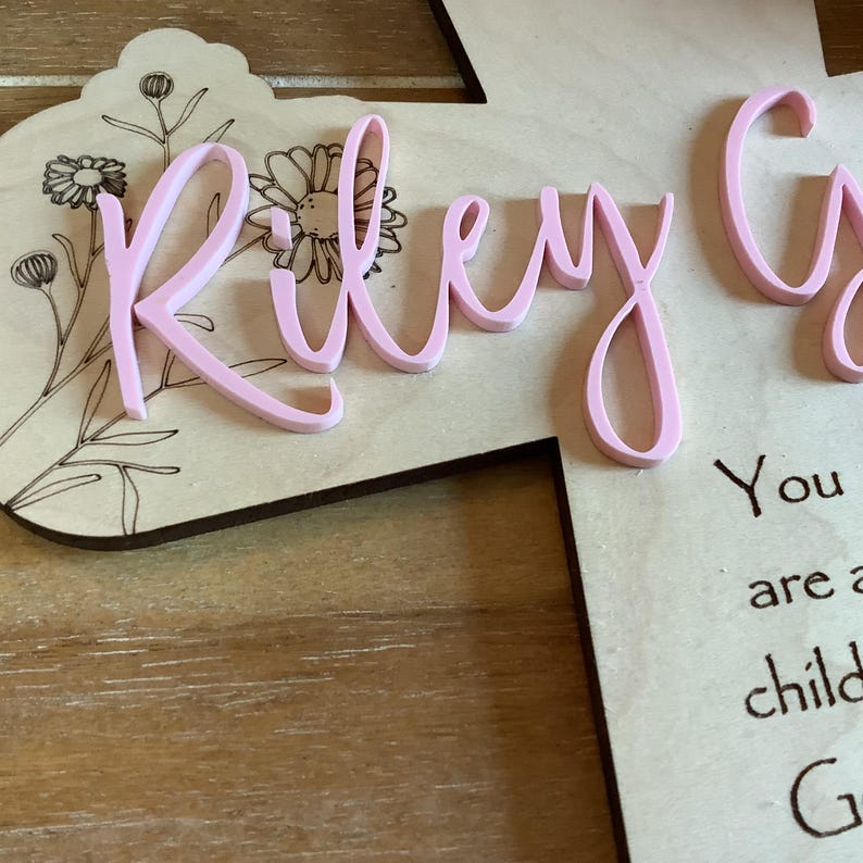 May include: A light-coloured wooden plaque featuring laser-engraved floral line art and the name Riley in raised, light pink script lettering. Below the name, black text is partially visible, reading You are a child G.