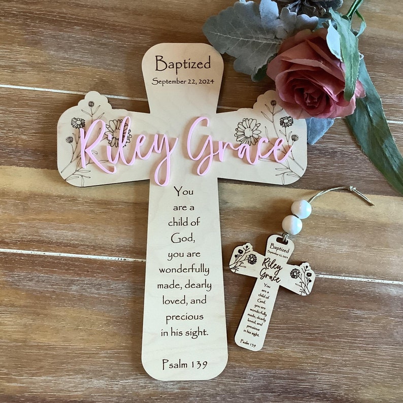 May include: A light-coloured wooden cross featuring the engraved text "Baptized September 22, 2024" and "Psalm 139" with a religious verse, accented by floral line art and the name "Riley Grace" in raised pink script lettering, alongside a smaller matching cross ornament.