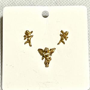 May include: Three gold-colored angel charms on a white card. Two angels are in a running pose, and one is in a flying pose. The charms are small and detailed, with wings and flowing garments. The card has a small hole at the top.
