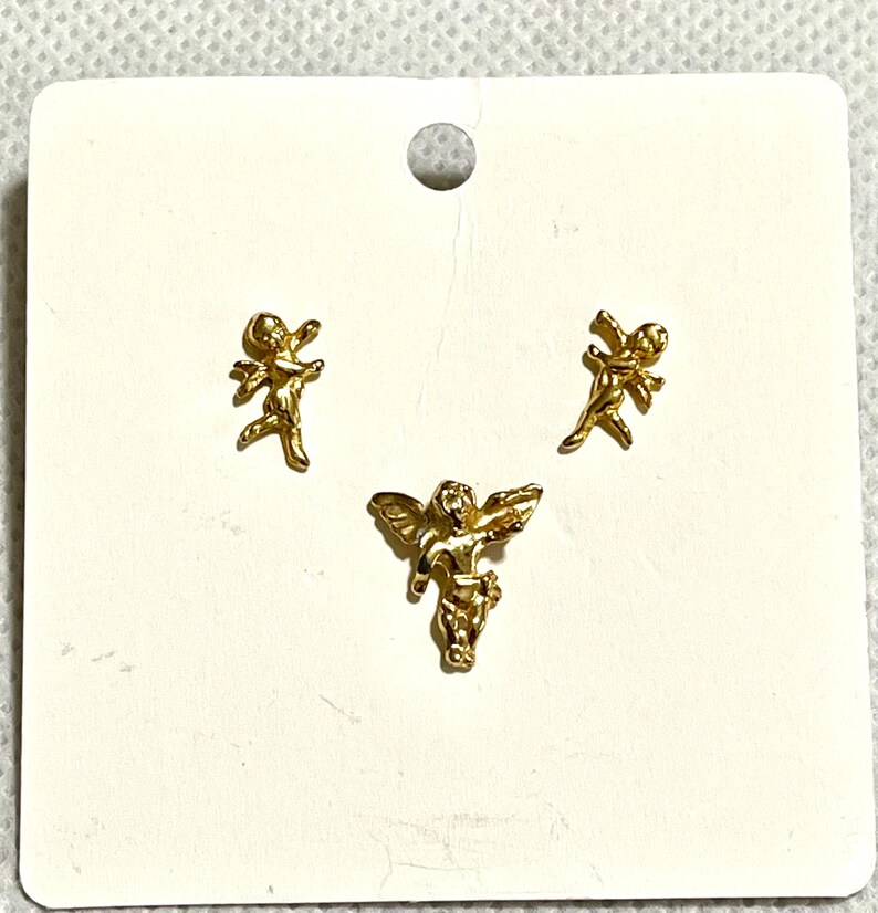 May include: Three gold-colored angel charms on a white card. Two angels are in a running pose, and one is in a flying pose. The charms are small and detailed, with wings and flowing garments. The card has a small hole at the top.