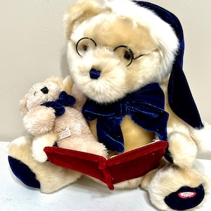 May include: A plush teddy bear wearing round glasses, a blue bow tie, and a matching hat. The bear holds a red book and a smaller bear. The bears are cream-colored with blue and red accents. The small bear has a blue bow tie.