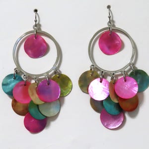 Może przedstawiać: Para kolczyków z wiszącymi, srebrnymi obręczami i skupiskiem kolorowych, opalizujących, okrągłych zawieszek. Zawieszki są w odcieniach różu, turkusu, zieleni i brązu. Kolczyki mają przyciągać wzrok i dodawać koloru.