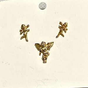 May include: Three gold-colored angel-shaped charms. Two are small, and one is larger with outstretched wings. The charms are on a light-colored background, possibly for jewelry making or crafting.