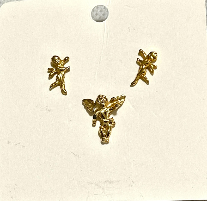 May include: Three gold-colored angel-shaped charms. Two are small, and one is larger with outstretched wings. The charms are on a light-colored background, possibly for jewelry making or crafting.