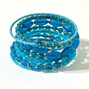 May include: A multi-strand wrap bracelet featuring a variety of blue and turquoise beads in different shapes and sizes, including oval, round, and faceted glass beads, interspersed with small metallic silver spacer beads on a flexible wire frame.